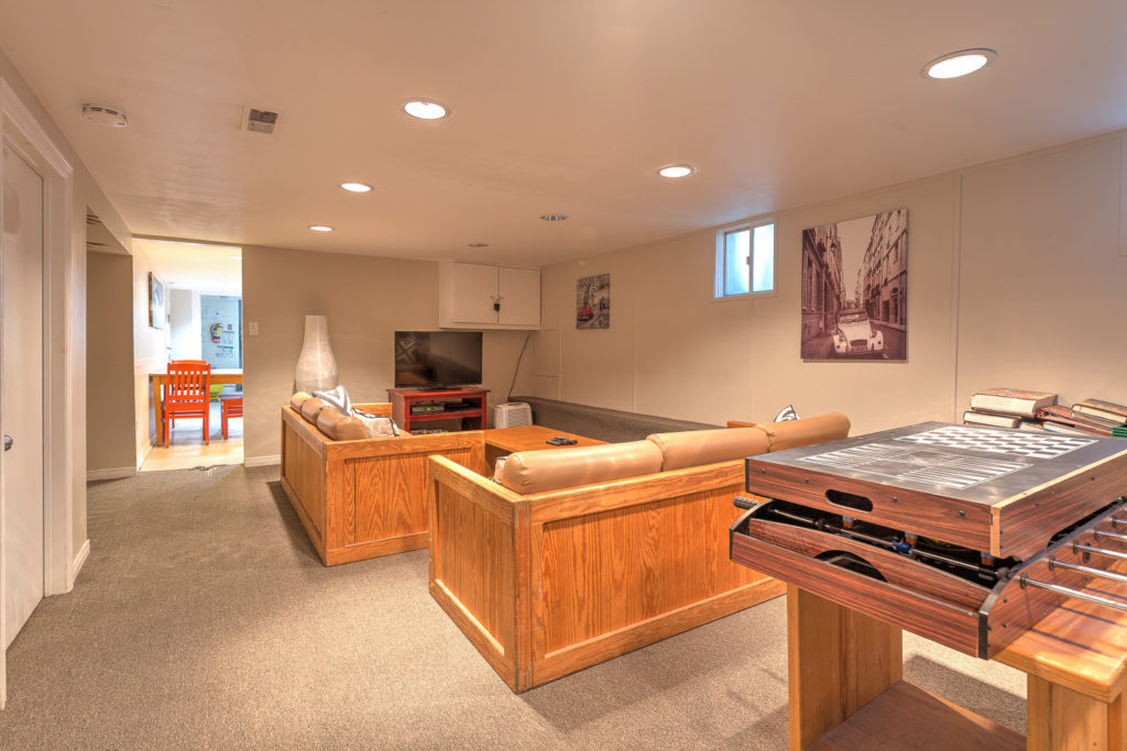 Interior shot of 106 Redwood Avenue. A recreation room with a foosball table with a chess board occupy the foreground, with two couches and a TV in the background. An opening into the dining area occupies the back.